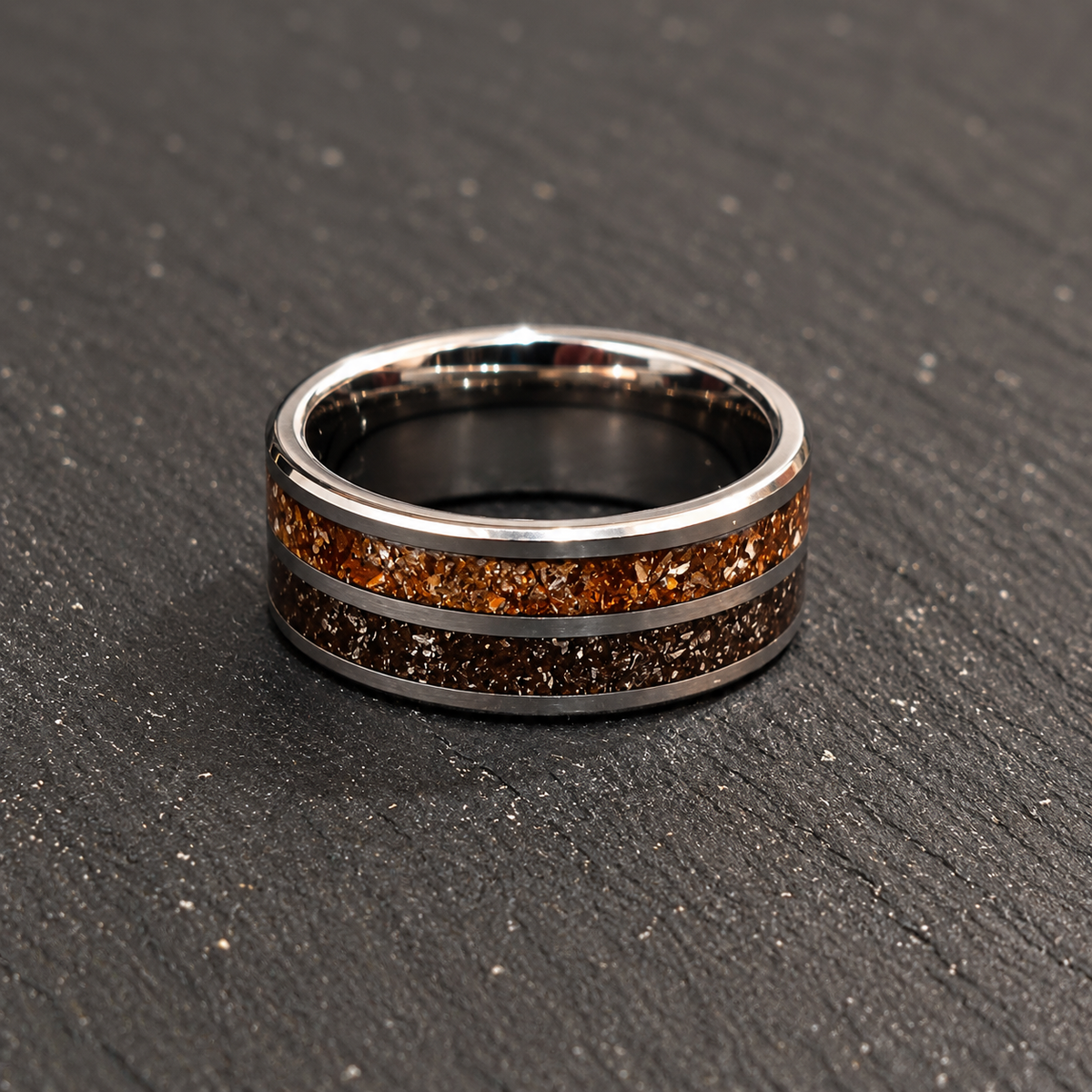 A polished silver-toned band ring with two textured inlay stripes, one amber-orange and one dark brown, resting on a dark gray textured surface.