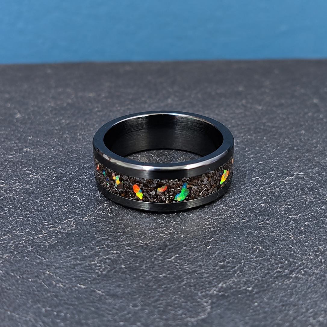 A black metal ring with a textured inlay of dark gravel and small multicolor opal flecks, displayed on a dark gray textured surface with a blue background.