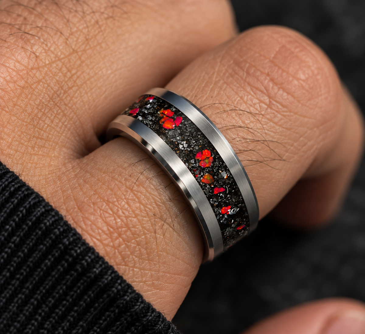 A close-up of a silver-toned ring worn on a finger, featuring a textured black inlay with scattered red and orange flecks.