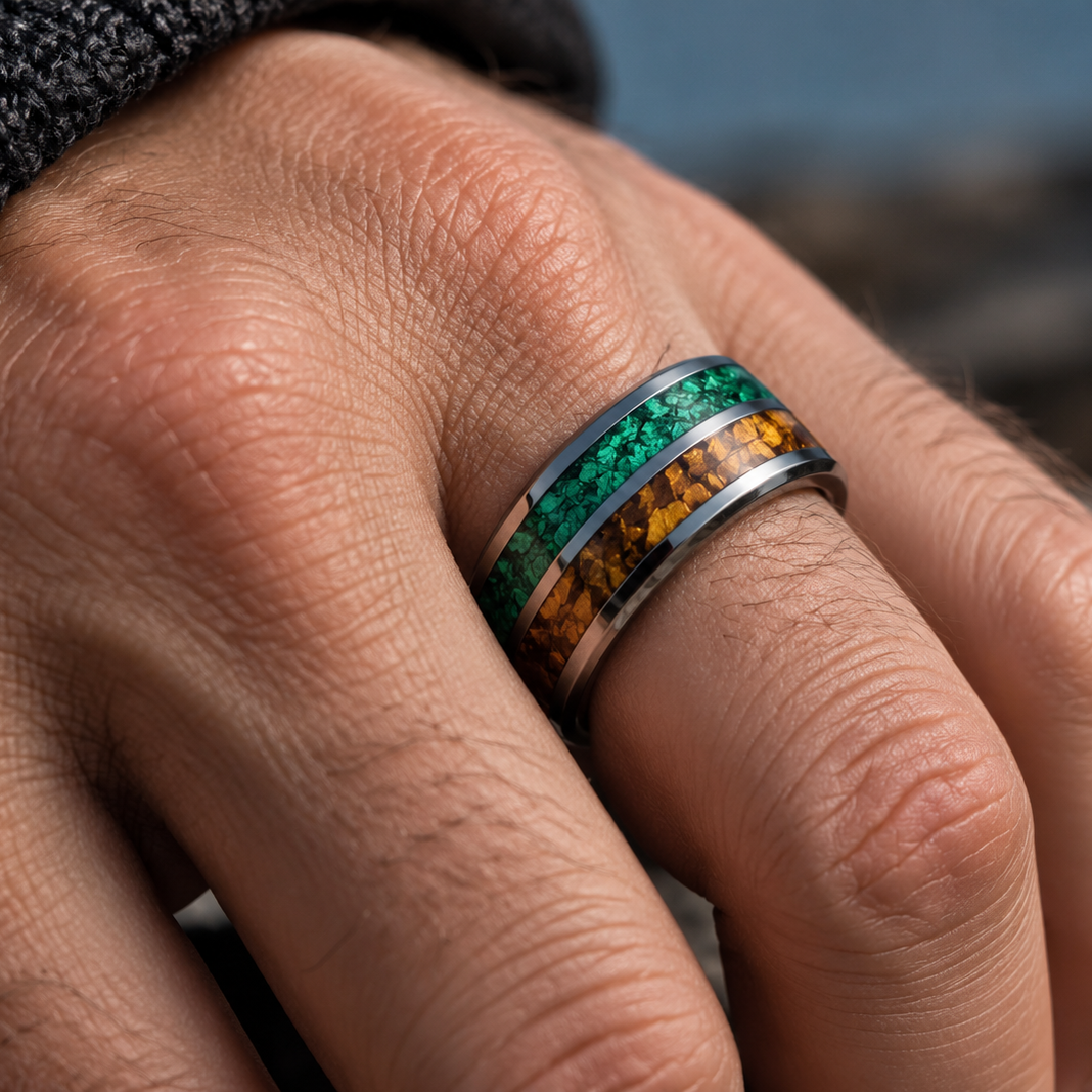 A close-up of a hand wearing a silver band ring with a green inlay on one side and a textured orange-brown inlay on the other, set against a blurred outdoor background.