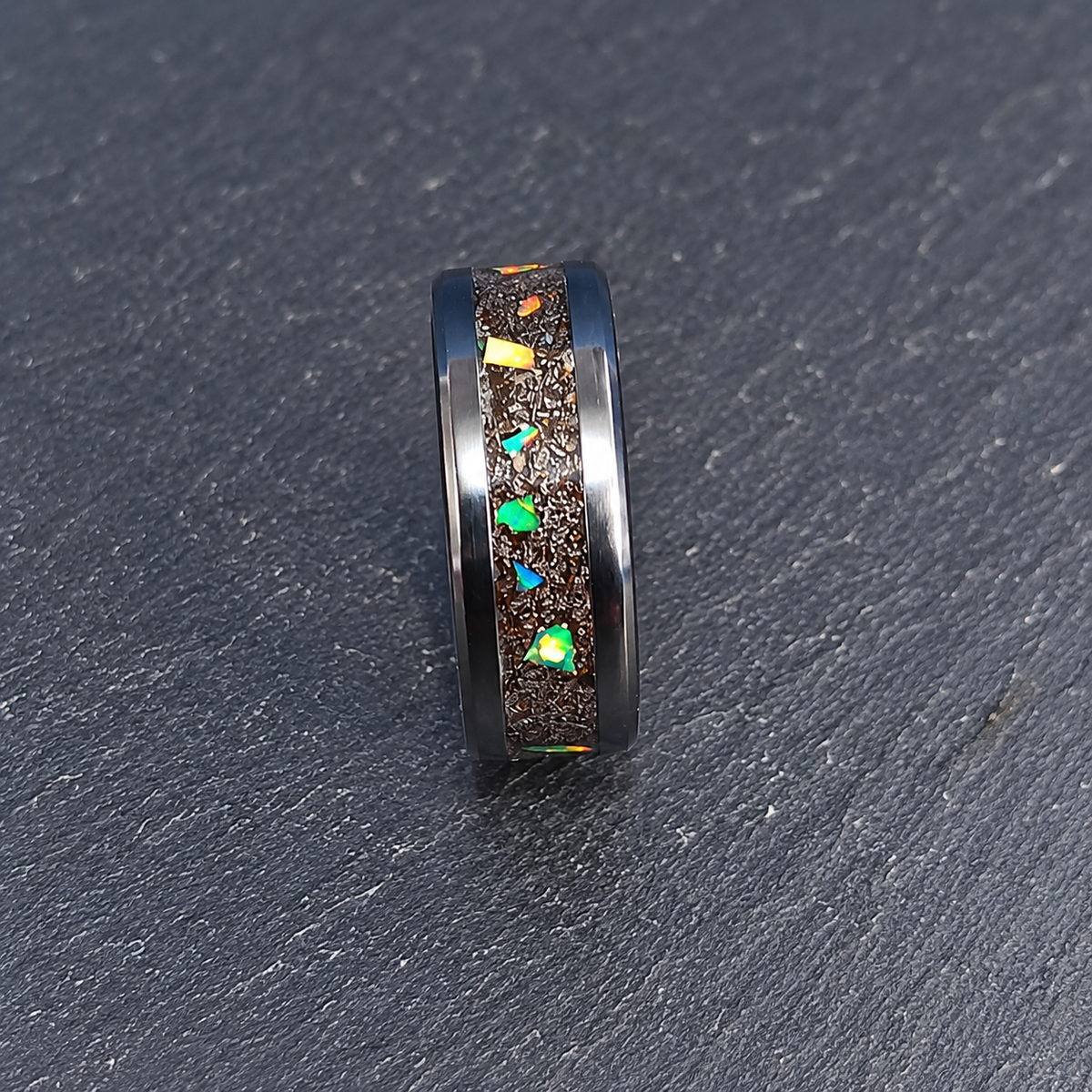 A black band ring with a textured brown inlay featuring colorful iridescent opal-like chips, shown on a dark textured slate background.