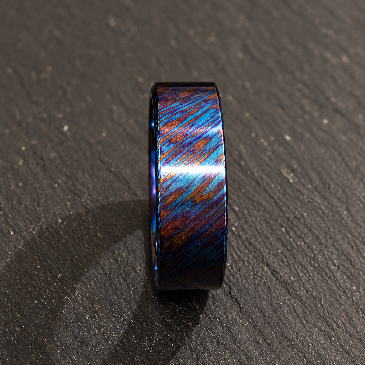 A black metallic ring with a vibrant blue, purple, and orange iridescent inlay, standing upright on a dark textured slate surface.