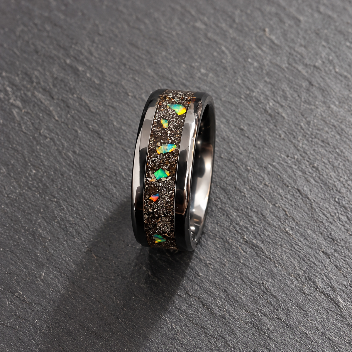 A polished dark metallic ring with a textured inlay of small multicolored opal-like stones, photographed on a dark gray stone surface.