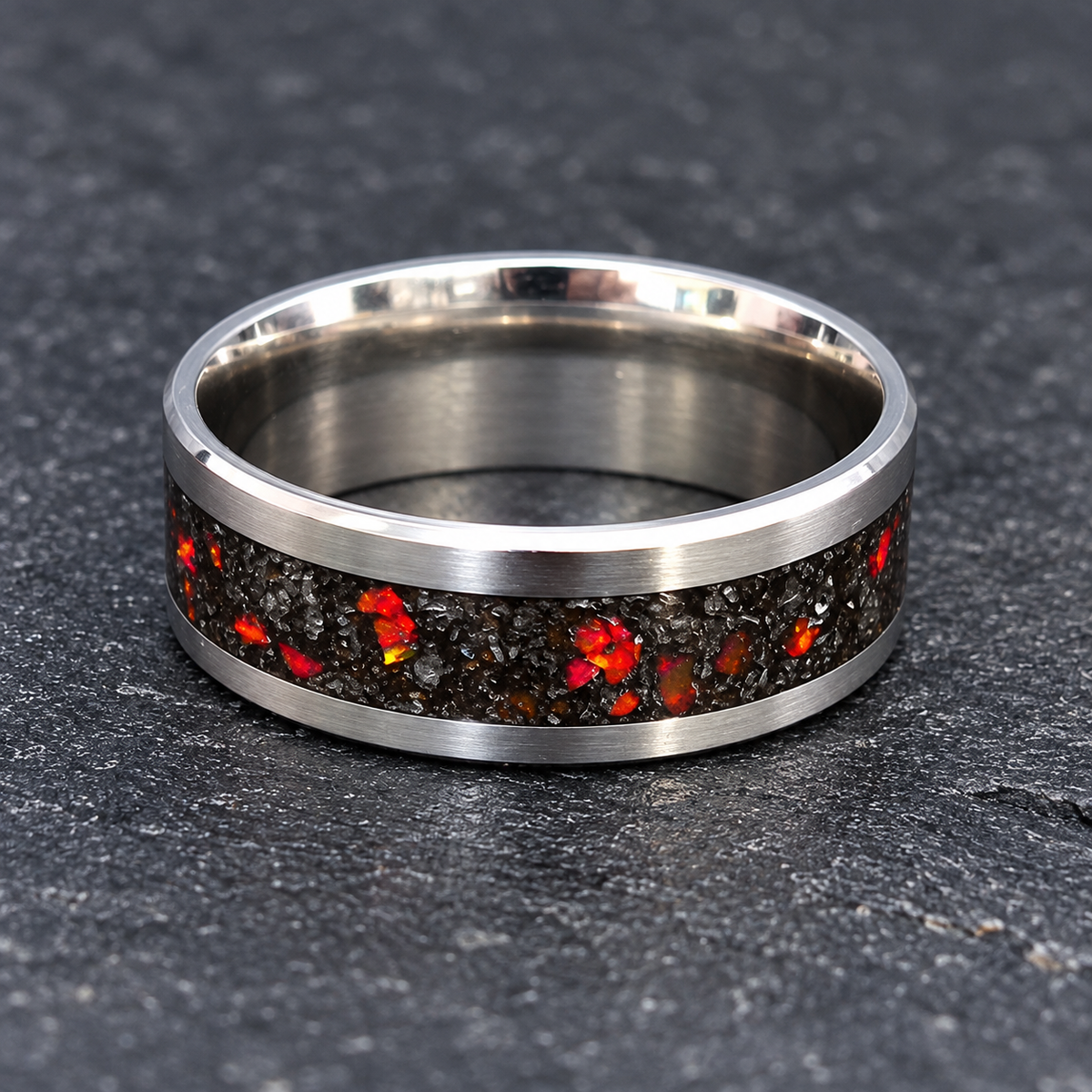 A silver-toned metal ring band with a brushed finish and a central inlay of dark textured material featuring bright red, orange, and yellow fiery flecks, shown on a dark gray textured stone background.