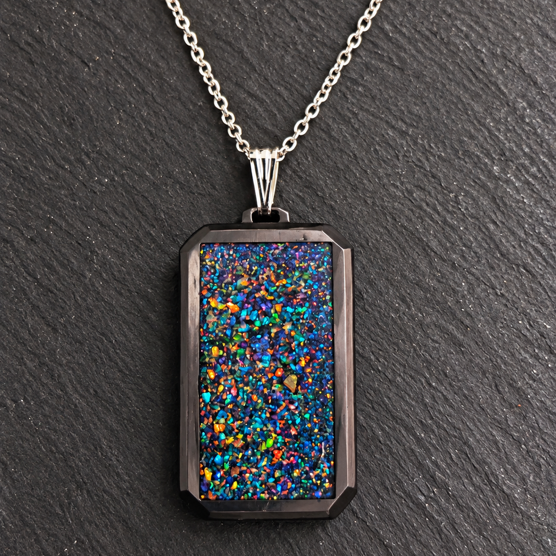 A silver-toned chain necklace with a rectangular pendant featuring a black background filled with dense, iridescent multicolor glitter, displayed on a dark slate surface.