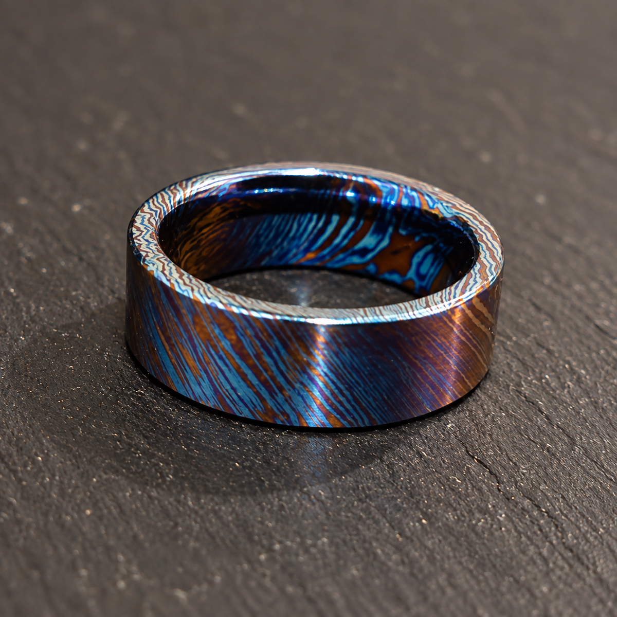A metallic ring band with a blue and copper iridescent pattern, resting on a dark textured slate surface.