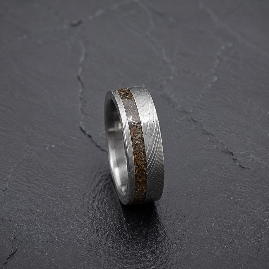A silver-toned metal ring with a textured wavy pattern and a central inlay of brown Dinosaur Bone, displayed on a dark textured slate surface.