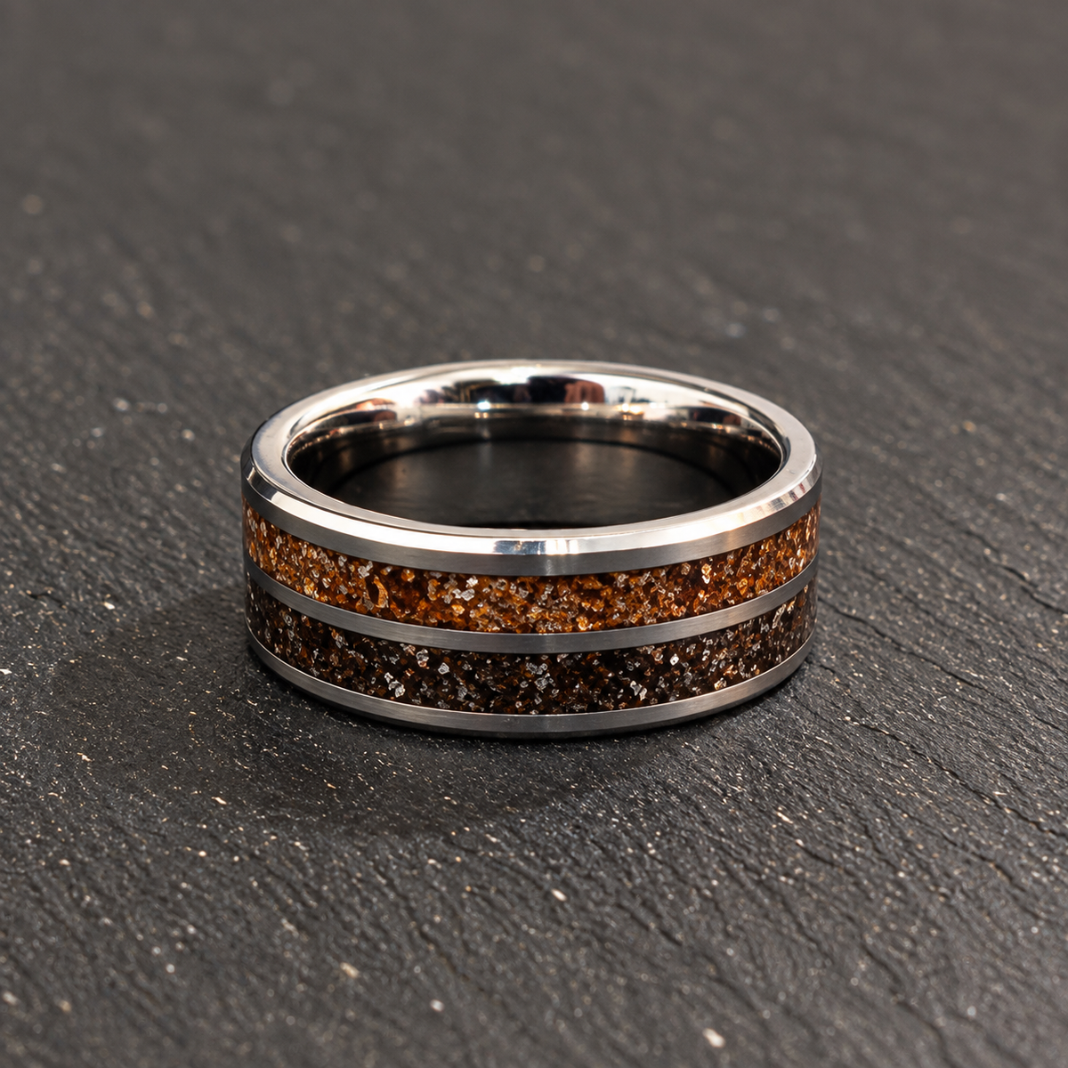 A polished silver-toned band ring with two inlay channels: one filled with amber-brown Dinosaur bone and the other with dark meteorite shavings, resting on a dark textured slate surface.