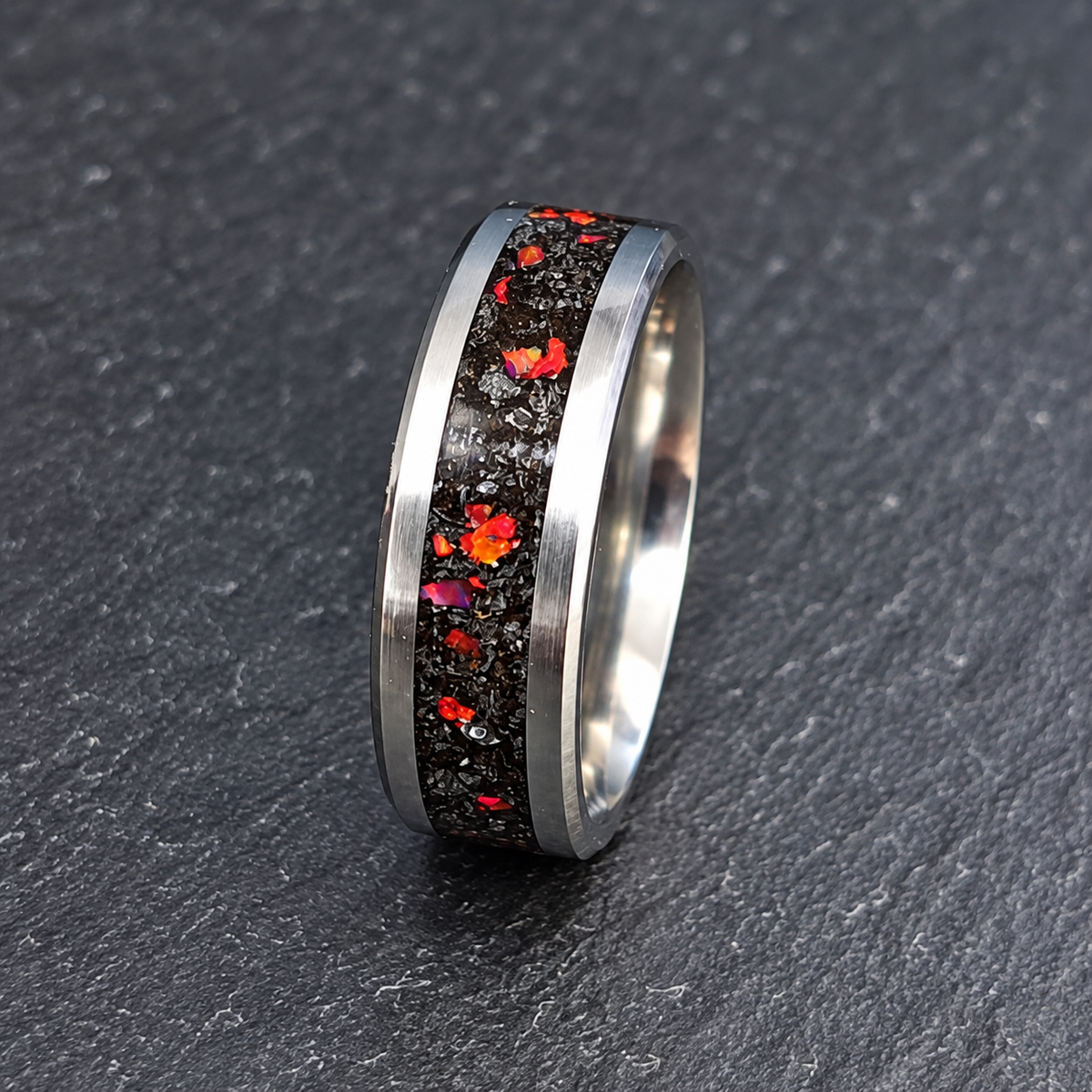 A silver-toned ring with a textured black inlay featuring scattered red, orange, and pink flecks, photographed on a dark gray textured surface.
