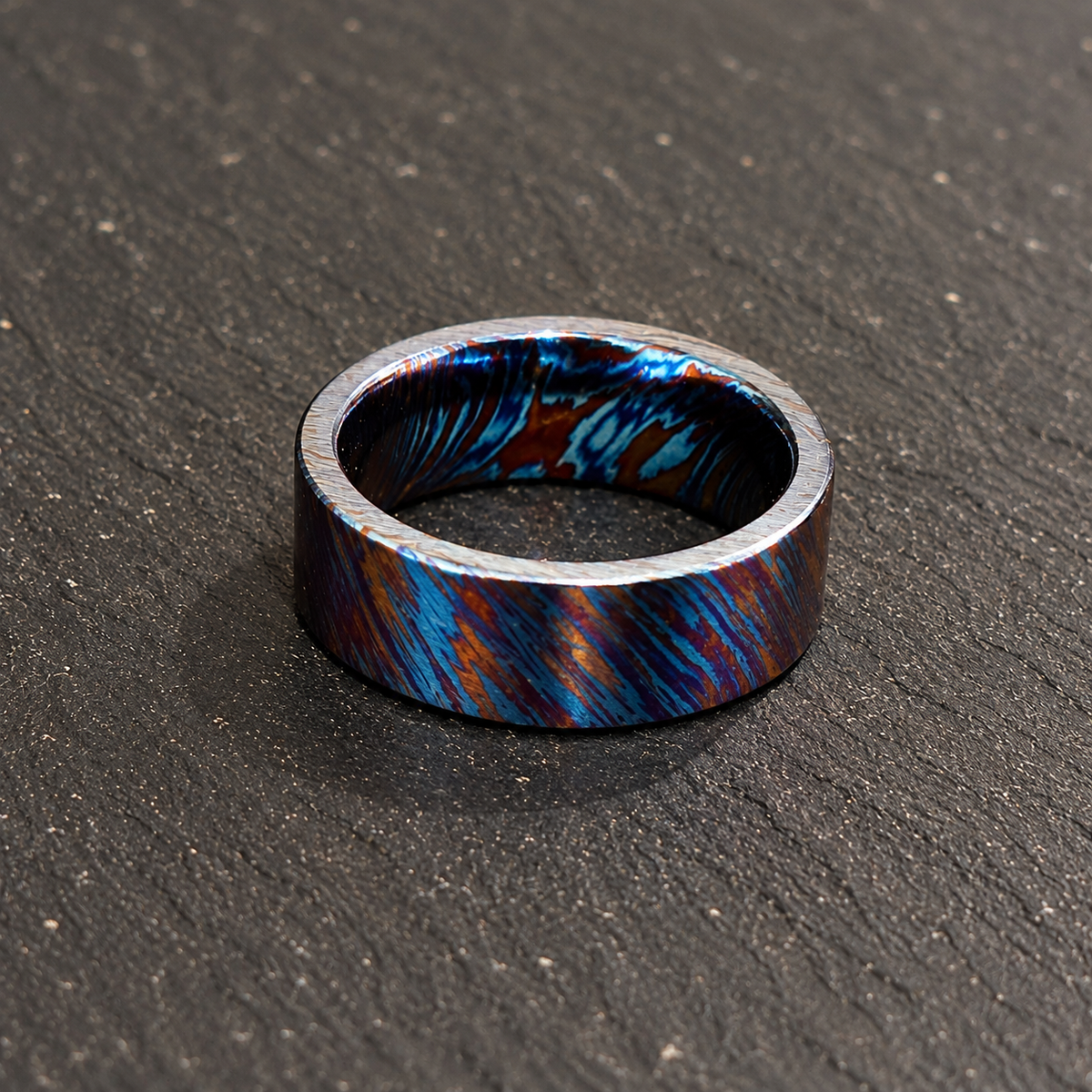 A metallic ring band with a marbled pattern of vibrant blue, copper-orange, and dark gray, resting on a dark textured slate surface.