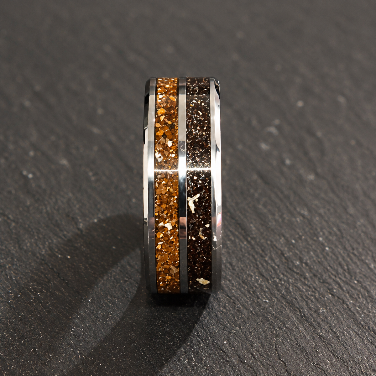 A polished silver-toned ring with two textured inlay channels, one filled with metallic gold flakes and the other with dark brown flakes, resting on a dark gray slate surface.