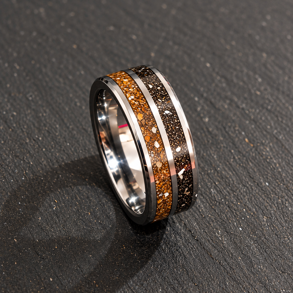 A polished silver-toned ring with a dual inlay design, featuring a textured orange-brown and black cosmic glitter band on the outer face, resting on a dark textured slate surface.