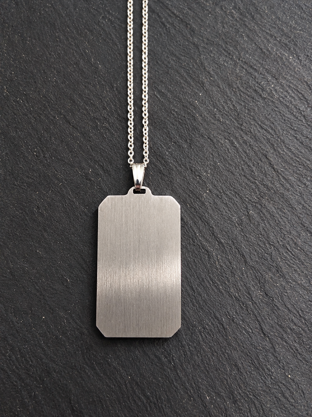 A brushed silver metal dog tag pendant necklace on a delicate silver chain, displayed against a dark textured slate background.