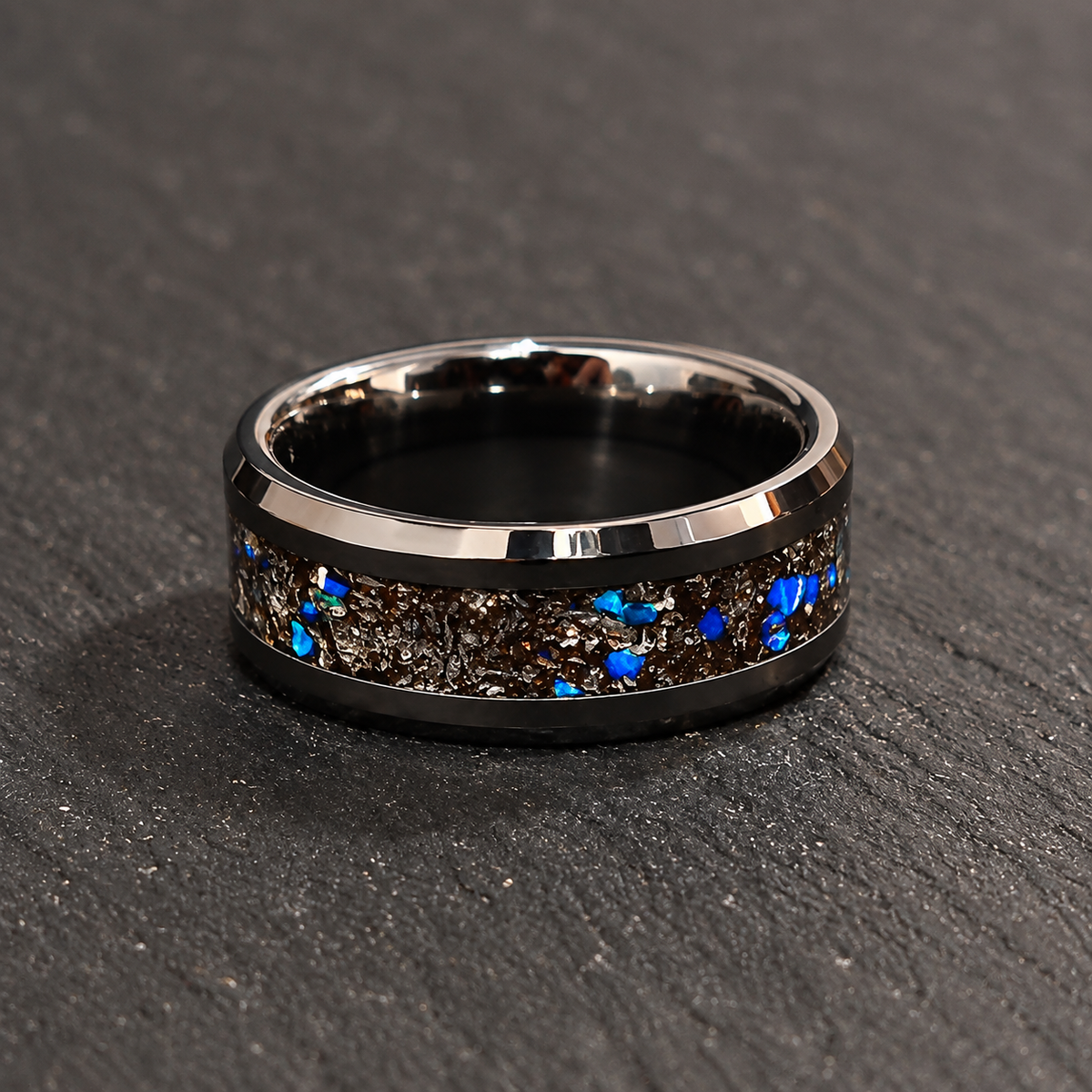 A polished black band ring with a textured inlay of metallic silver flakes and bright blue iridescent chips, resting on a dark gray slate surface.