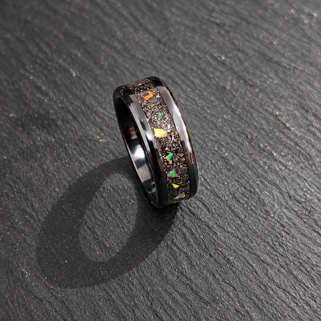 A black metallic ring featuring a textured inlay of small multicolored iridescent flecks, photographed on a dark slate surface.