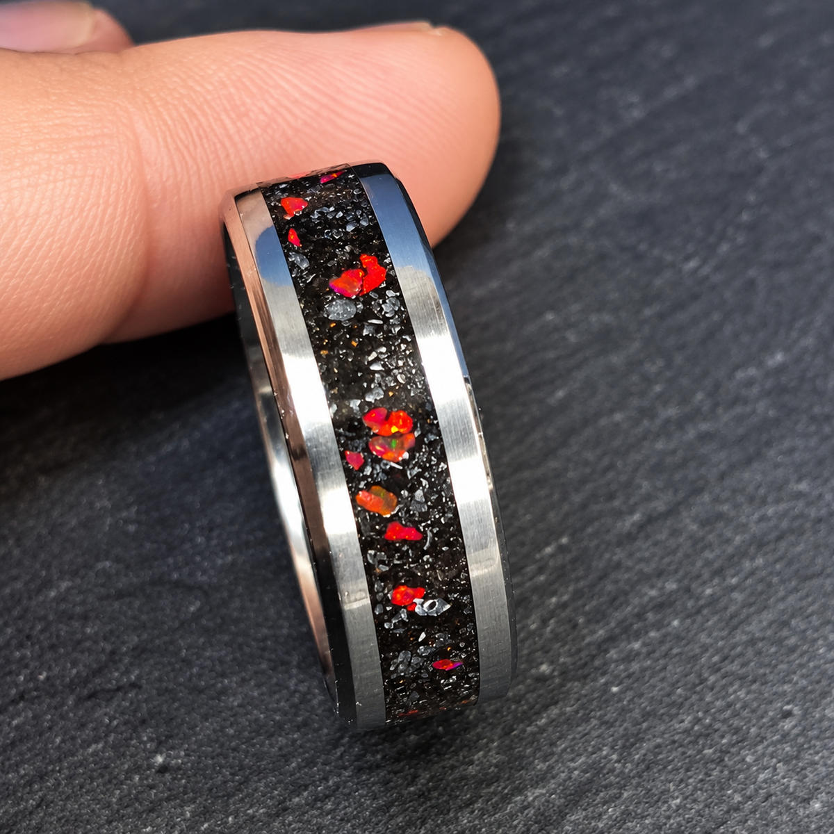 A close-up of a silver-toned ring with a black inlay containing red and orange flecks, held between fingers on a dark textured surface.