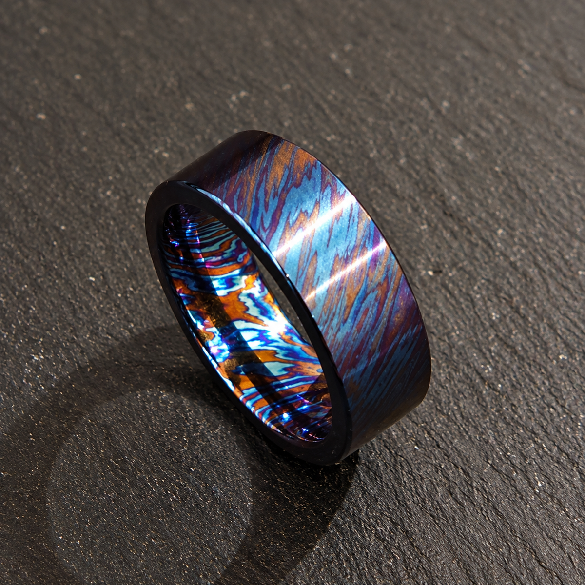 A metallic ring band with a dark blue and orange marbled or swirled pattern, photographed on a dark textured surface.