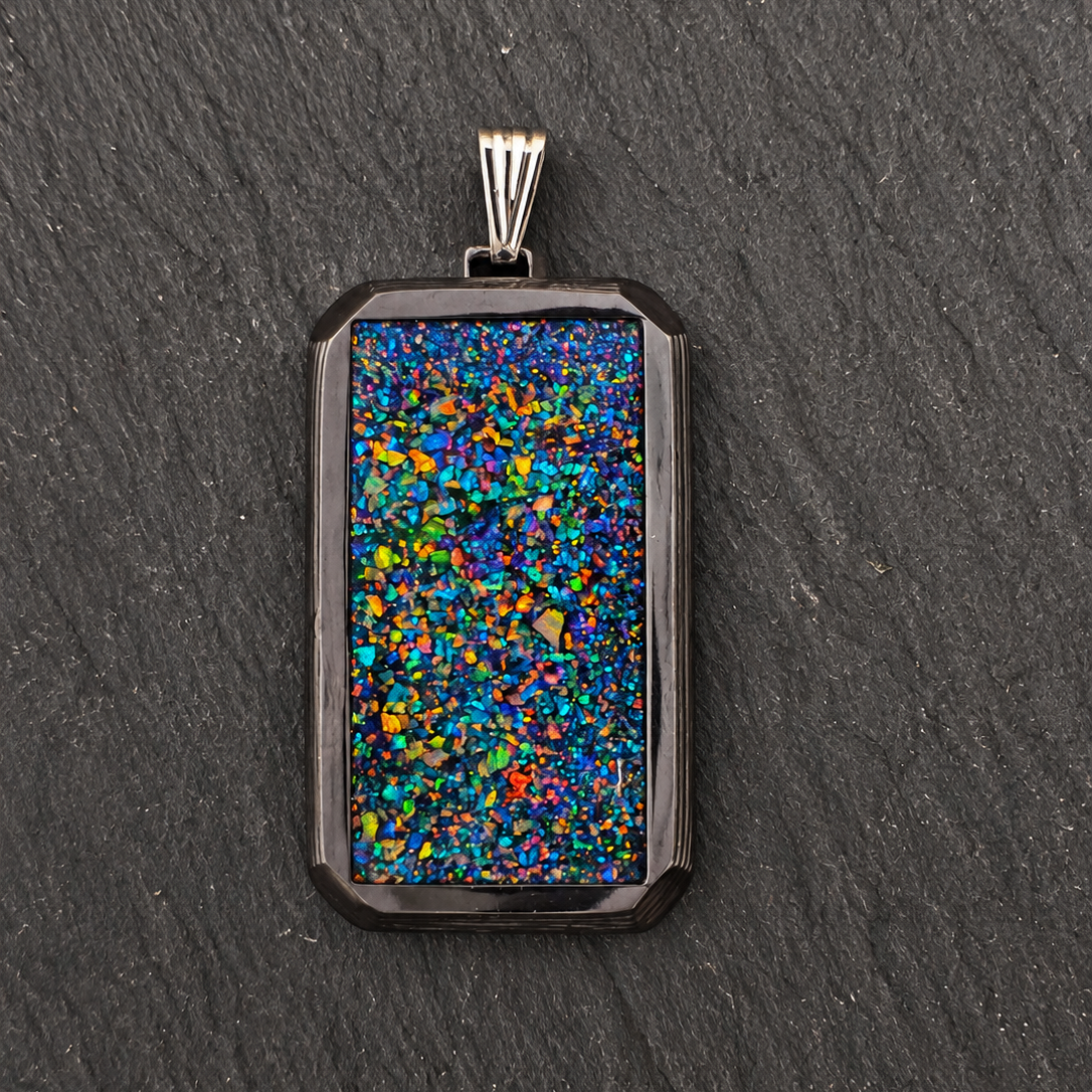 A rectangular pendant with a black bezel and a vibrant, multicolored opal inlay, resting on a dark gray slate background.