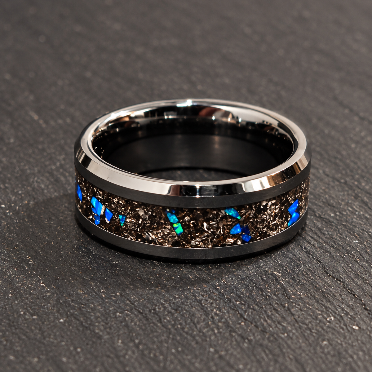 A polished silver-toned band ring with a black inlay featuring metallic gold flecks and small vibrant blue opal-like inclusions, displayed on a dark textured slate surface.
