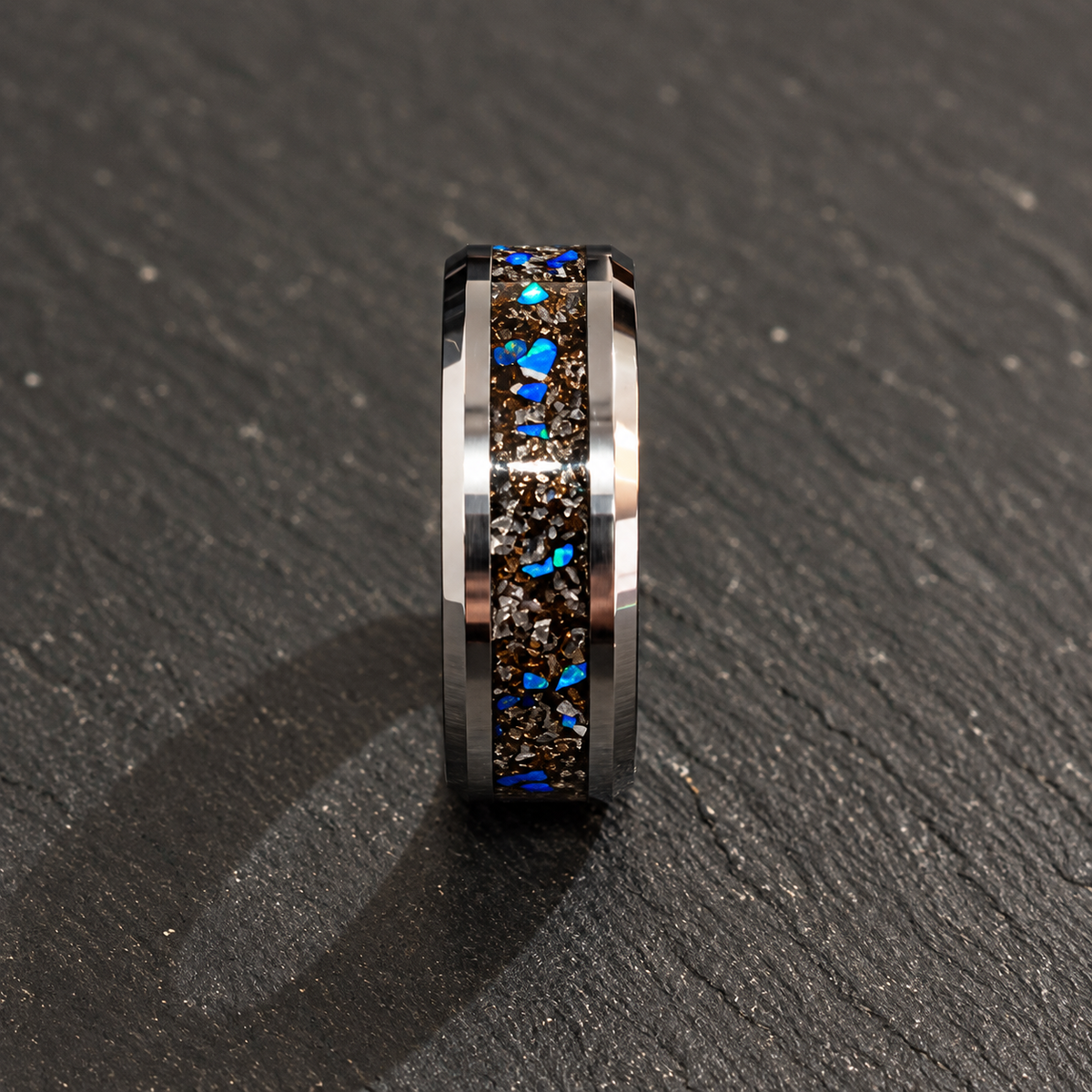 A polished silver-toned ring with a central inlay of dark brown and black mineral chips and bright blue opal-like flecks, photographed standing upright on a dark textured surface.