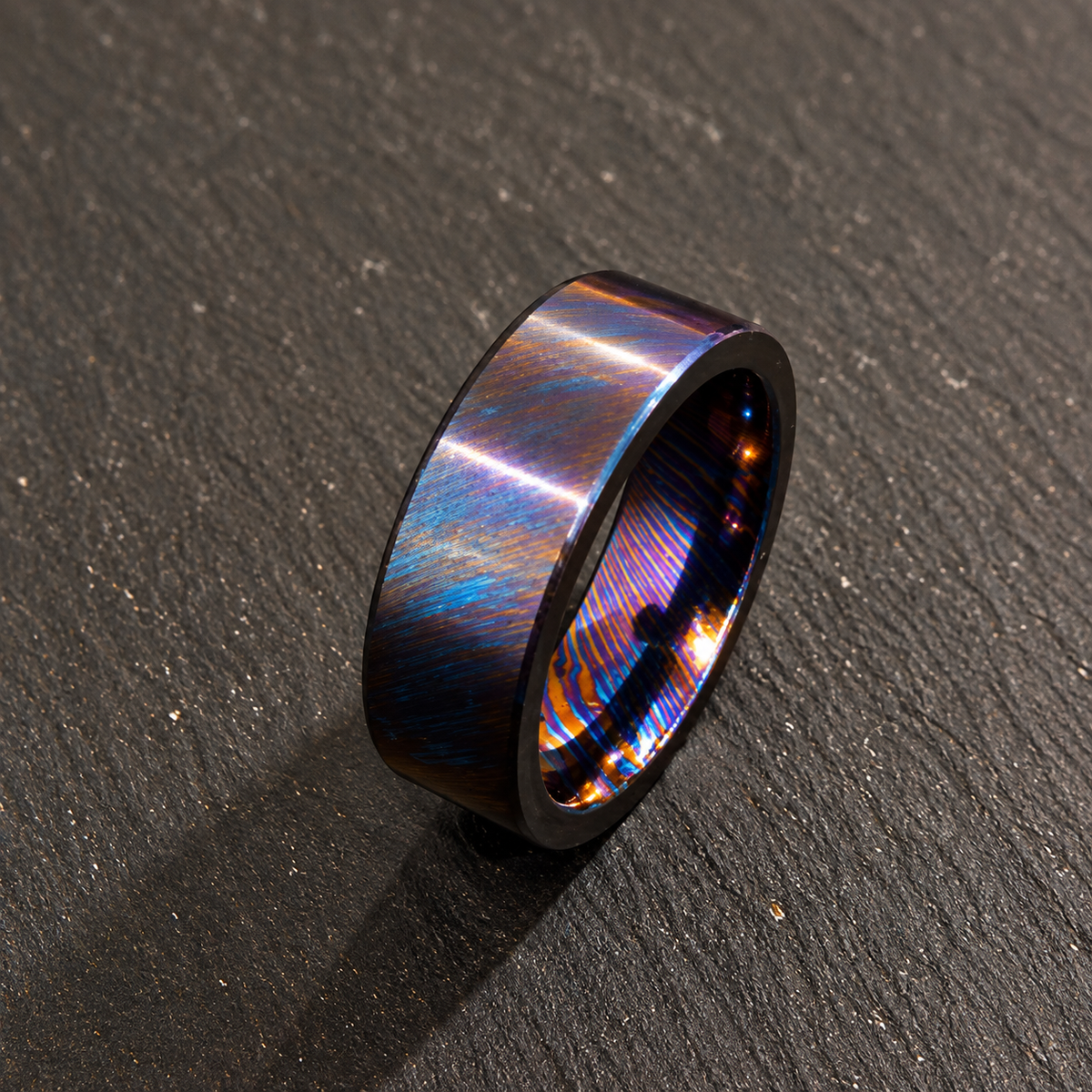 A dark metallic ring with a vibrant iridescent blue, purple, and orange flame-patterned inlay, resting on a dark textured slate surface.