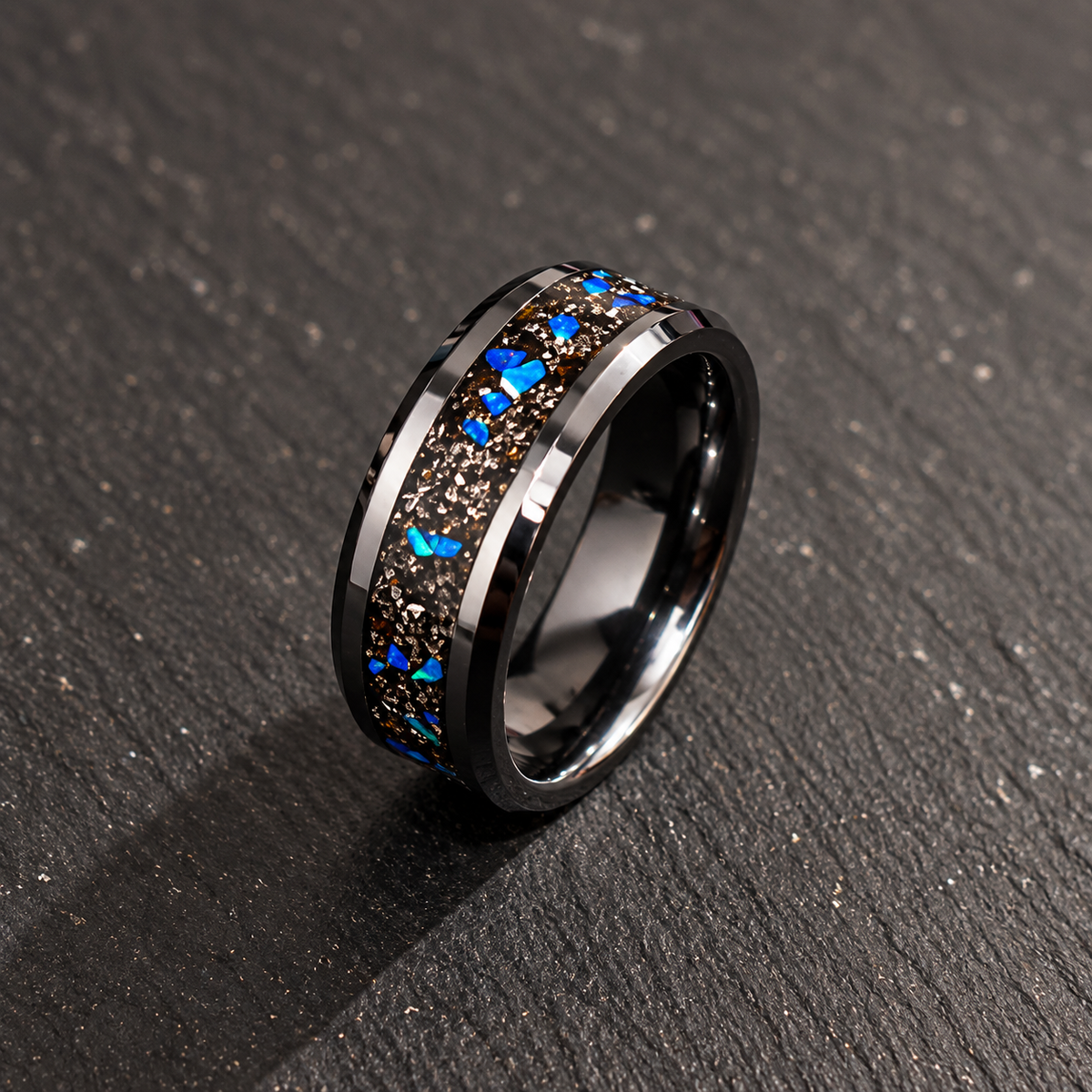 A polished black metal ring band with a textured dark brown inlay and bright blue glitter accents, resting on a dark textured surface.
