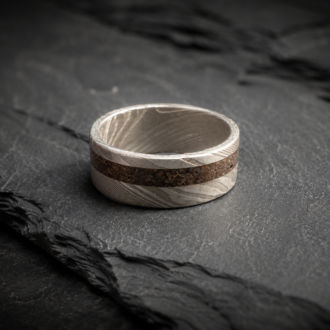 A silver-toned ring with a textured wood grain pattern and a central brown inlay, resting on a dark slate surface.