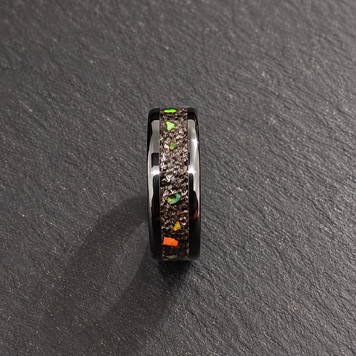 A black ring with a textured, metallic silver inlay featuring small colorful flecks, photographed on a dark gray slate surface.