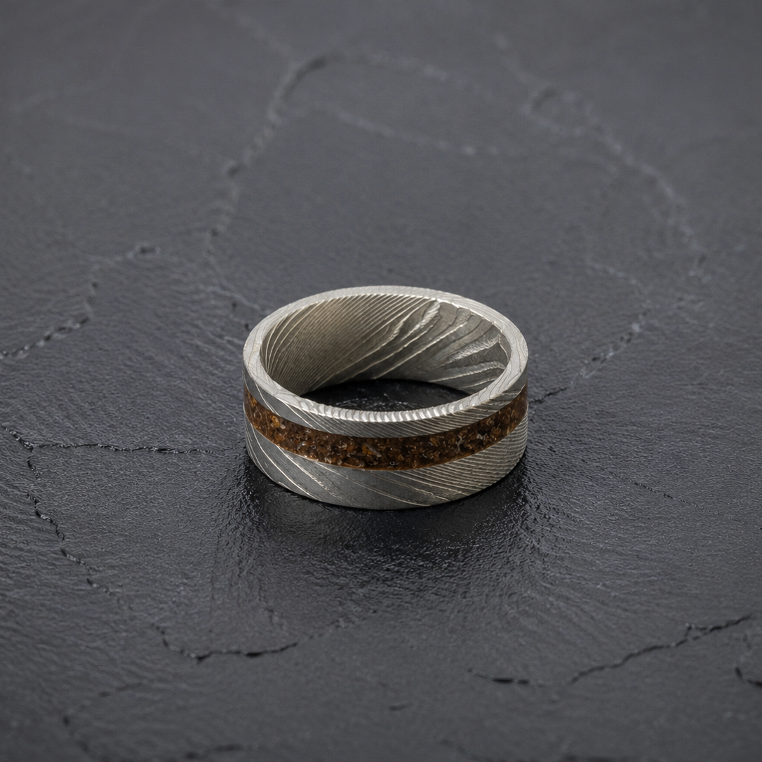 A silver-toned ring with a textured, feather-like pattern and a central brown inlay, displayed on a dark textured slate surface.