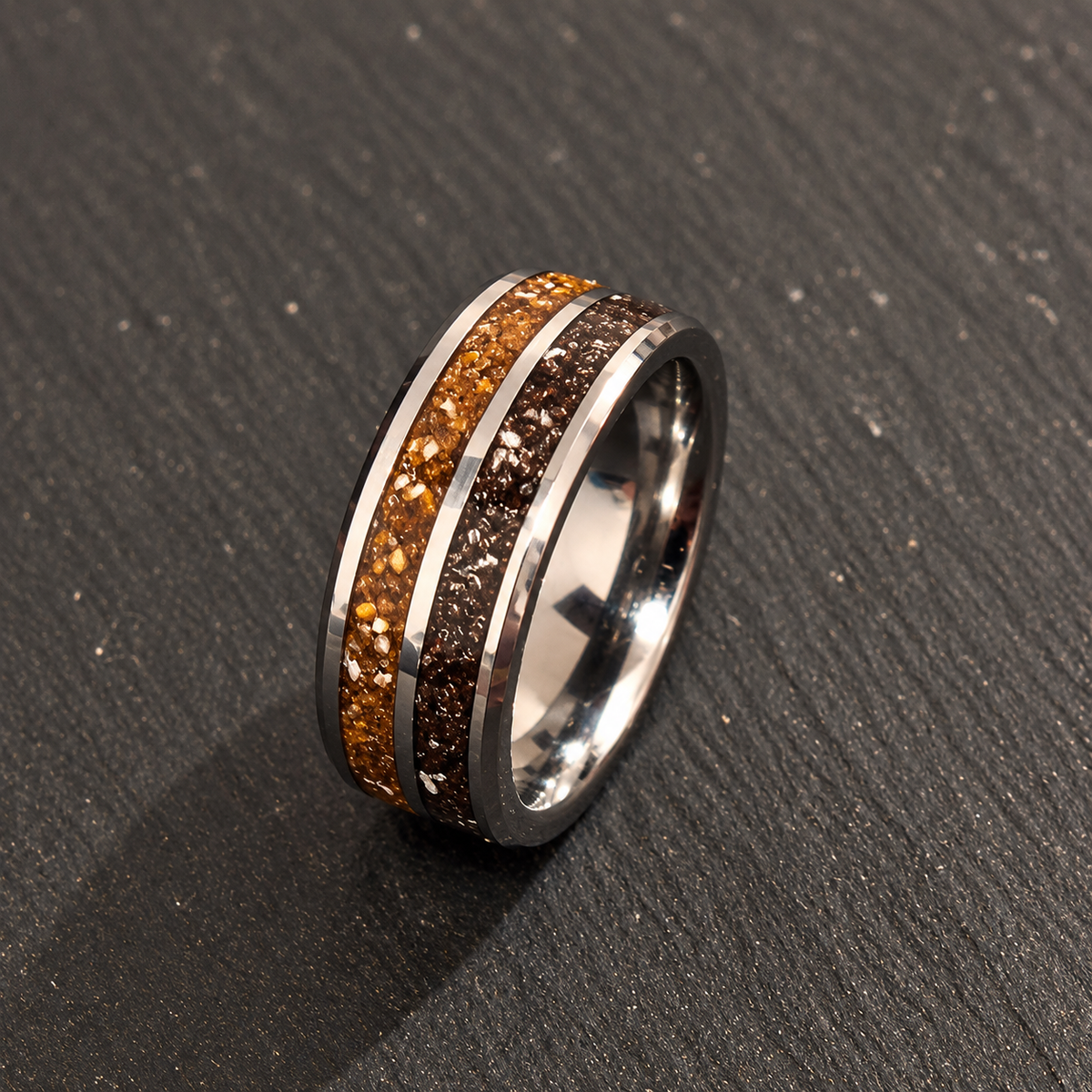 A polished silver-toned band ring with two inlay channels: one filled with golden-brown glitter and the other with dark brown glitter, resting on a dark textured slate surface.